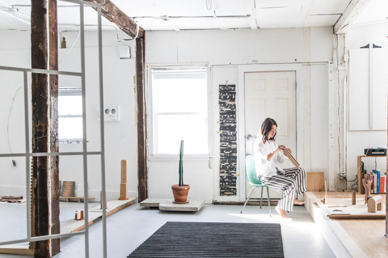 An woman artist sitting in the corner of a white room as she works with a small wooden sculpture.