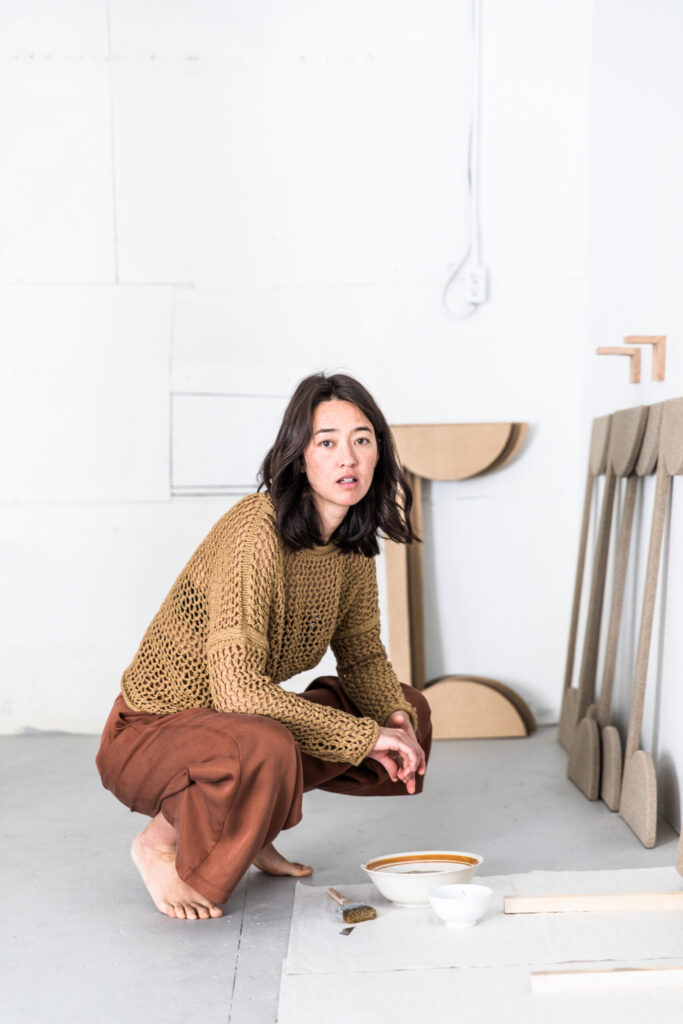 A woman artist is captured in a white studio as she is crouched down over a bowl of paint and wooden shapes are leaning against the wall.