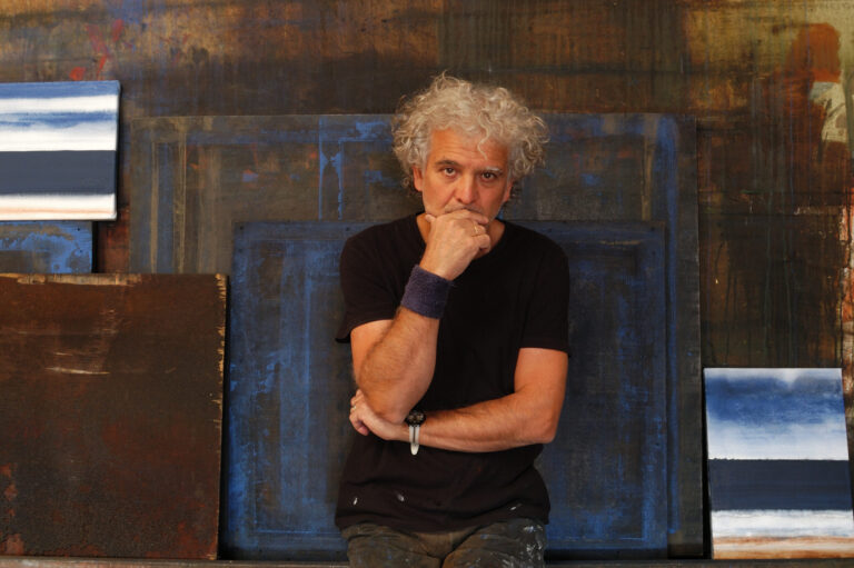 A male artist with white and wild hair poses with his hand to his mouth in front of several dark paintings.