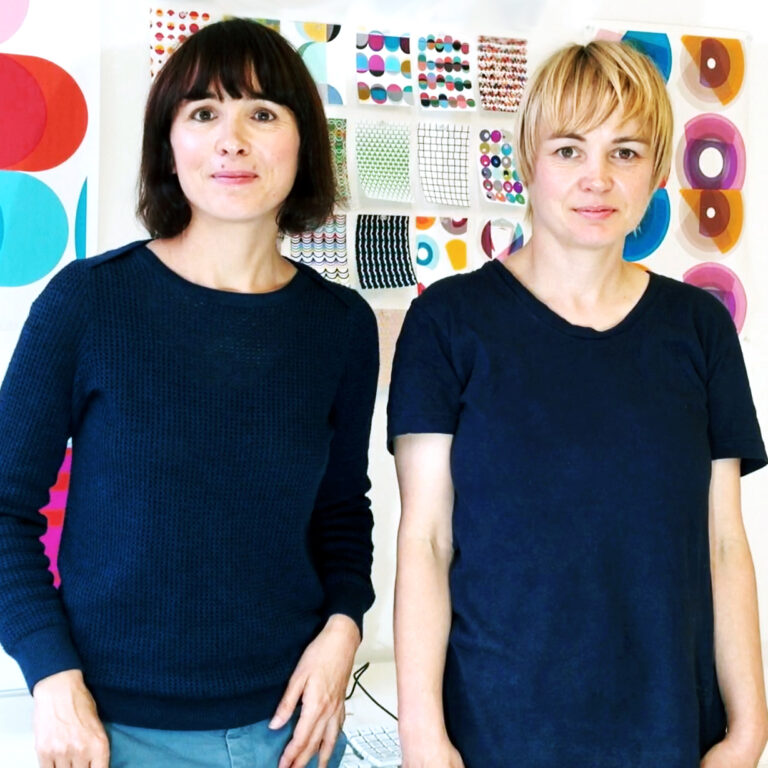 A pair of woman artist, one blonde and one with dark hair pose for a portait in front of several colorful and geometric concepts pinned to the wall behind them.