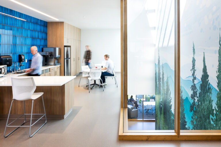 An office kitchen with a couple individuals present. The kitchen has a large glass wall that looks out onto a wall featuring a forest design.